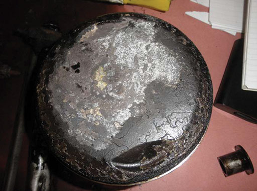 Photo 8: The no. 4 piston head of the No. 3 Engine, after it had been removed by investigators during the engine teardown. Note the whitish coloring and the damaged texture, indicative of detonation due to premature ignition.